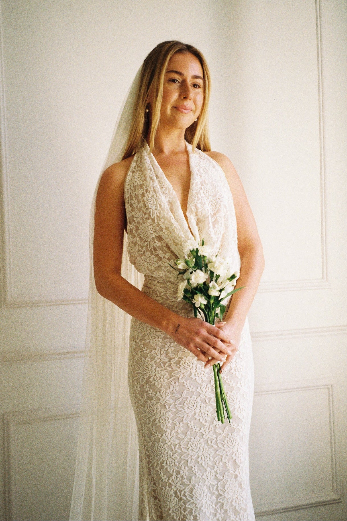 Woman in a white lace dress holding a bouquet against a white paneled wall.